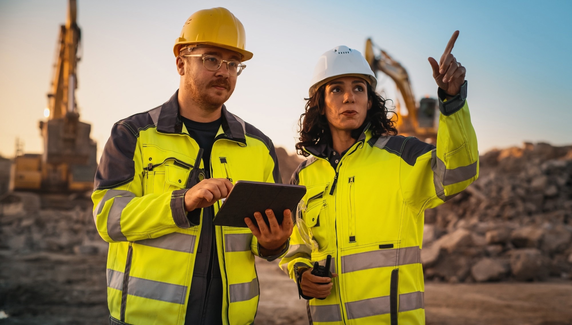 A male and female mining worker in hi-vis. The man holds a screen, while the woman points something out in the distance.