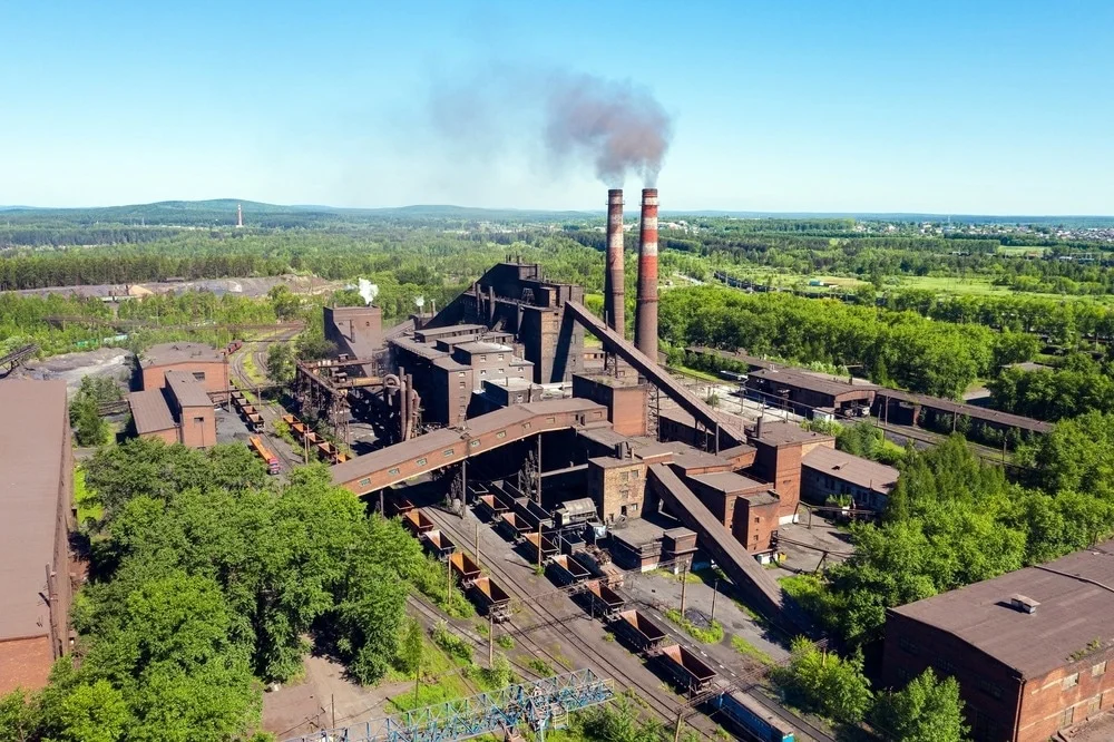 An image of a manufacturing plant in surrounded by trees etc. with smoke billowing from two large chimneys.